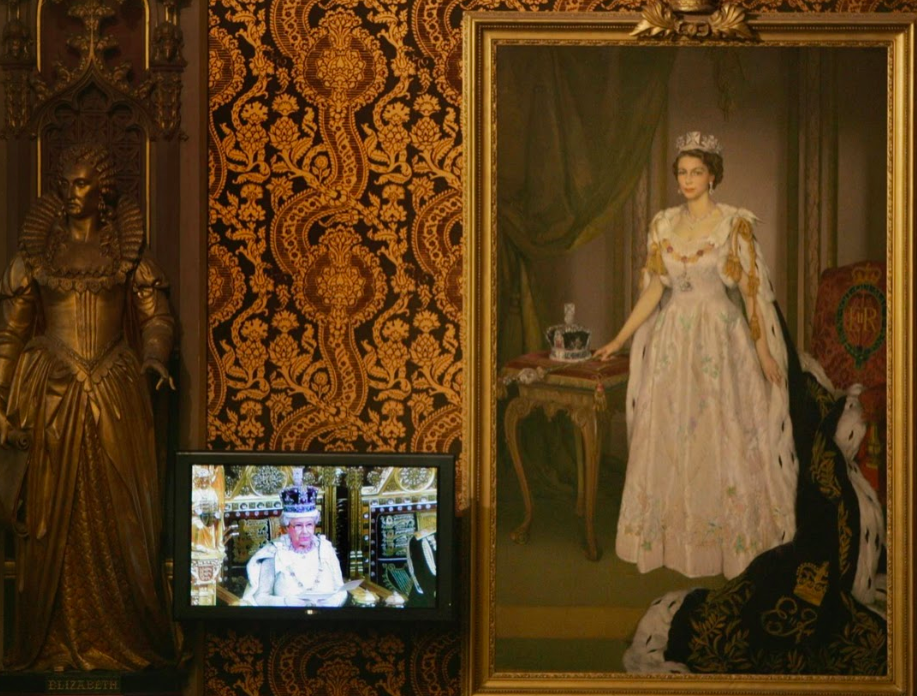 الملكة تظهر على شاشة التلفاز، إلى جوار لوحةٍ لها، في أثناء إلقائها خطاباً بافتتاح دورة البرلمان.